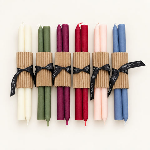 Six double taper candles in assorted colors wrapped in brown corrugated paper bands and tied with black ribbons, arranged on a light wood surface.