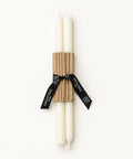 A set of two white taper candles wrapped in a brown corrugated paper sleeve and tied with a black ribbon printed with the brand name 'Candle Box', placed on a light wood surface.