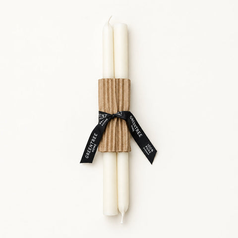 A set of two white taper candles wrapped in a brown corrugated paper sleeve and tied with a black ribbon printed with the brand name 'Candle Box', placed on a light wood surface.