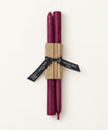 Two dark purple candles wrapped in a brown corrugated paper sleeve and tied with a black ribbon printed with the brand name 'The Entree', placed on a light wood background.