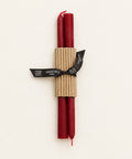 Two red taper candles wrapped in a corrugated kraft paper sleeve and tied with a black ribbon label that reads 'GREENTREE WOOD', placed on a light wood surface.