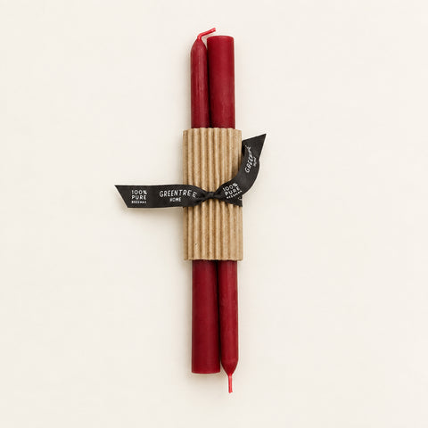 Two red taper candles wrapped in a corrugated kraft paper sleeve and tied with a black ribbon label that reads 'GREENTREE WOOD', placed on a light wood surface.