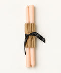 Two pale pink taper candles wrapped together in a brown corrugated paper sleeve and tied with a black ribbon printed with the brand name GREENE, placed on a warm wood surface.