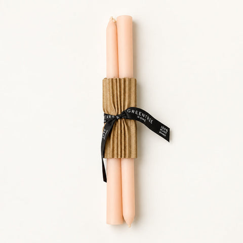 Two pale pink taper candles wrapped together in a brown corrugated paper sleeve and tied with a black ribbon printed with the brand name GREENE, placed on a warm wood surface.
