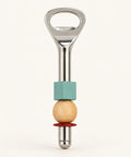 A bottle opener styled with a turquoise block, a light wood bead, and a red ring, resting on a light wood grain surface.