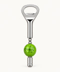 A novelty bottle opener with a polished silver metal body and a bright green spherical charm featuring a black line-art flower design, shown on a light wood grain background.