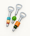 Three bottle openers arranged on a light wood tabletop. Each opener has a silver metal loop and handle with decorative beads and cylindrical accents in turquoise, natural wood, green, orange, and dark green.