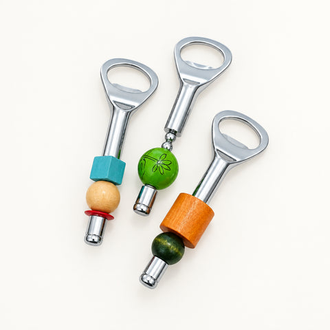Three bottle openers arranged on a light wood tabletop. Each opener has a silver metal loop and handle with decorative beads and cylindrical accents in turquoise, natural wood, green, orange, and dark green.