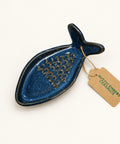 A dark blue-gray ceramic fish-shaped dish with a grater on top, resting on a wooden surface with a paper tag attached.