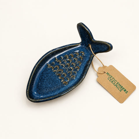 A dark blue-gray ceramic fish-shaped dish with a grater on top, resting on a wooden surface with a paper tag attached.
