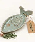 A fish-shaped ceramic grater with a pale sage green glaze and a small raised fish detail on the top, shown on a wooden tabletop next to a brown paper tag that reads 'Vestig Bakes Teaming'.