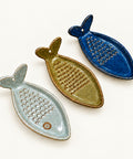 Three ceramic fish-shaped grater dishes in sage green, olive green, and dark blue, displayed on a light wood surface with handmade paper tags.