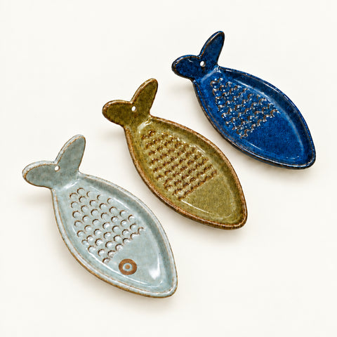 Three ceramic fish-shaped grater dishes in sage green, olive green, and dark blue, displayed on a light wood surface with handmade paper tags.