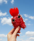 Red plush toy held by a hand against a blue sky with clouds