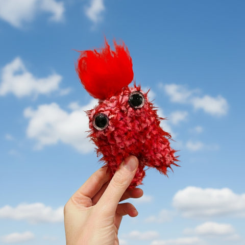 Red plush toy held by a hand against a blue sky with clouds