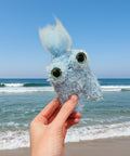 Hand holding a blue plush toy against a backdrop of clouds
