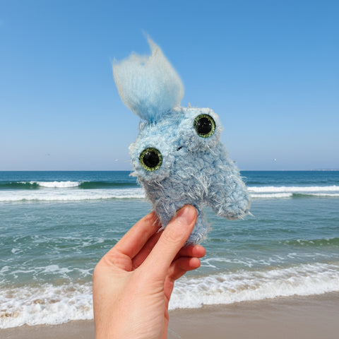 Hand holding a blue plush toy against a backdrop of clouds