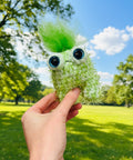 Hand holding a green plush toy with black eyes against a neutral background