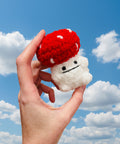 Hand holding a small plush toy with a red cap and white body against a plain background