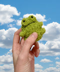 Hand holding a small green plush frog toy against a plain background