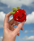 Hand holding a small red crocheted strawberry with a face against a plain background