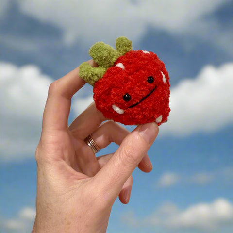Hand holding a small red crocheted strawberry with a face against a plain background