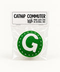 A green felt G train catnip toy sealed in a clear plastic bag with a white paper label that reads 'CATNIP COMMUTER' and 'MADE WITH LOVE IN ASTORIA, NYC', placed on a wooden surface.