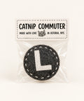 A circular grey felt L train catnip toy sealed in a clear plastic bag with a printed tag that reads 'CATNIP COMMUTER' on a wooden surface.