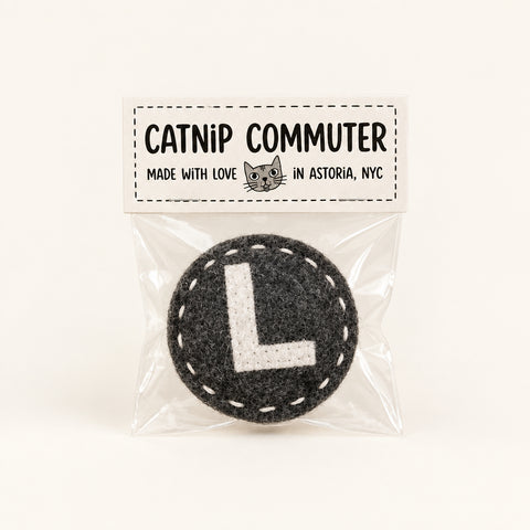 A circular grey felt L train catnip toy sealed in a clear plastic bag with a printed tag that reads 'CATNIP COMMUTER' on a wooden surface.