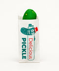 A green pickle in a white paper wrapper printed with a cartoon pickle and the words 'PICKLE Delicious', placed on a wooden surface.