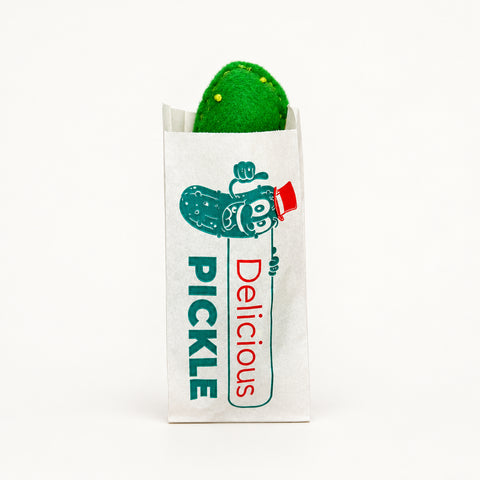 A green pickle in a white paper wrapper printed with a cartoon pickle and the words 'PICKLE Delicious', placed on a wooden surface.