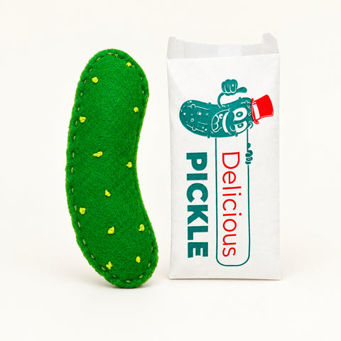 A green felt pickle-shaped item with yellow felt cplets and white stitching, placed next to a white paper bag printed with a cartoon pickle and the word 'PICKLE' on a light wood surface.