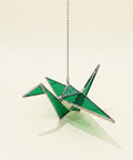 Green and silver crane-shaped ornament hanging on a chain over a wooden surface with a blurred background.