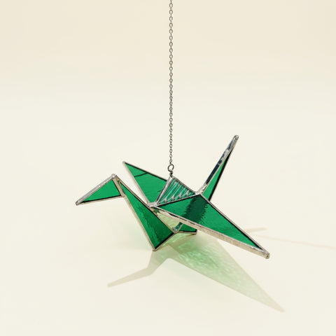 Green and silver crane-shaped ornament hanging on a chain over a wooden surface with a blurred background.