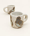 A ceramic mug with a speckled off-white glaze and a painted pigeon design, sitting on a light wood table with another mug blurred in the background.