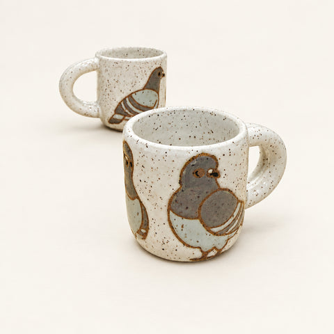 A ceramic mug with a speckled off-white glaze and a painted pigeon design, sitting on a light wood table with another mug blurred in the background.