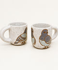 Two speckled ceramic mugs with painted pigeon designs, displayed on a wooden desk with a laptop in the background.