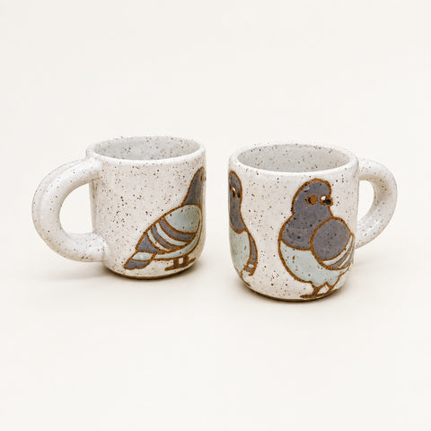 Two speckled ceramic mugs with painted pigeon designs, displayed on a wooden desk with a laptop in the background.