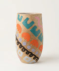 A tall, narrow ceramic vase with a gold glitter base and colorful abstract painted patterns, standing on a light wood desk in a shop setting.