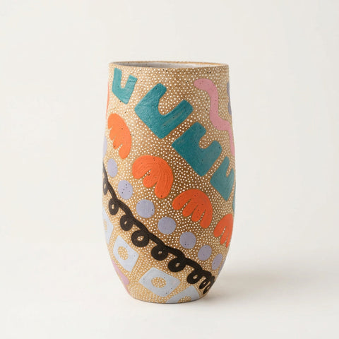 A tall, narrow ceramic vase with a gold glitter base and colorful abstract painted patterns, standing on a light wood desk in a shop setting.