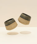 Two ceramic cups with speckled design and black accents on a beige background