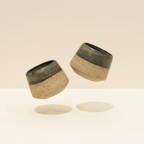 Two ceramic cups with speckled design and black accents on a beige background