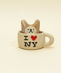 Small ceramic cat figurine inside a cup with 'I ❤️ NY' text on a wooden surface.