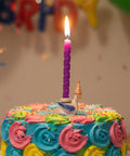 Colorful birthday cake with a lit candle in front of balloons and confetti.