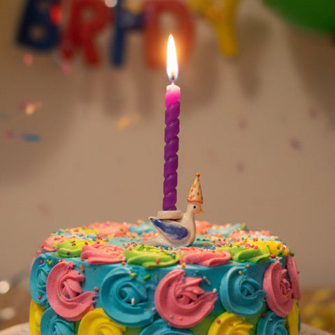 Colorful birthday cake with a lit candle in front of balloons and confetti.