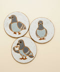 Three ceramic coasters with pigeon designs on a beige background