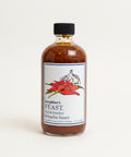 Bottle of Josephine's Feast hot and smoky Sriracha sauce on a wooden table with a blurred office background.