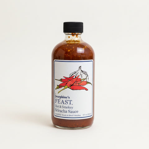 Bottle of Josephine's Feast hot and smoky Sriracha sauce on a wooden table with a blurred office background.