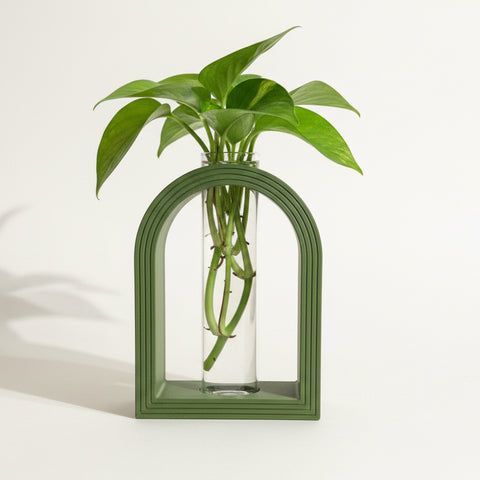 Green arch-shaped vase with a clear glass tube and plant trimmings inside of it.
