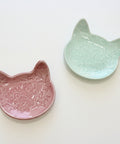 Two ceramic cat-shaped dishes, one pink and one green, on a wooden surface.